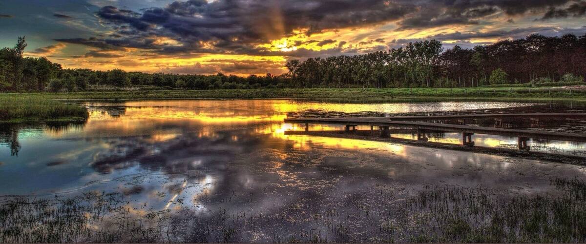 A sunset over a calm river with a wooden bridge and dense trees in the background.