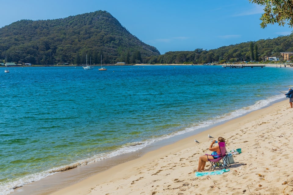 A beach with clear turquoise water, a sandy shore, and a forested hill in the background.