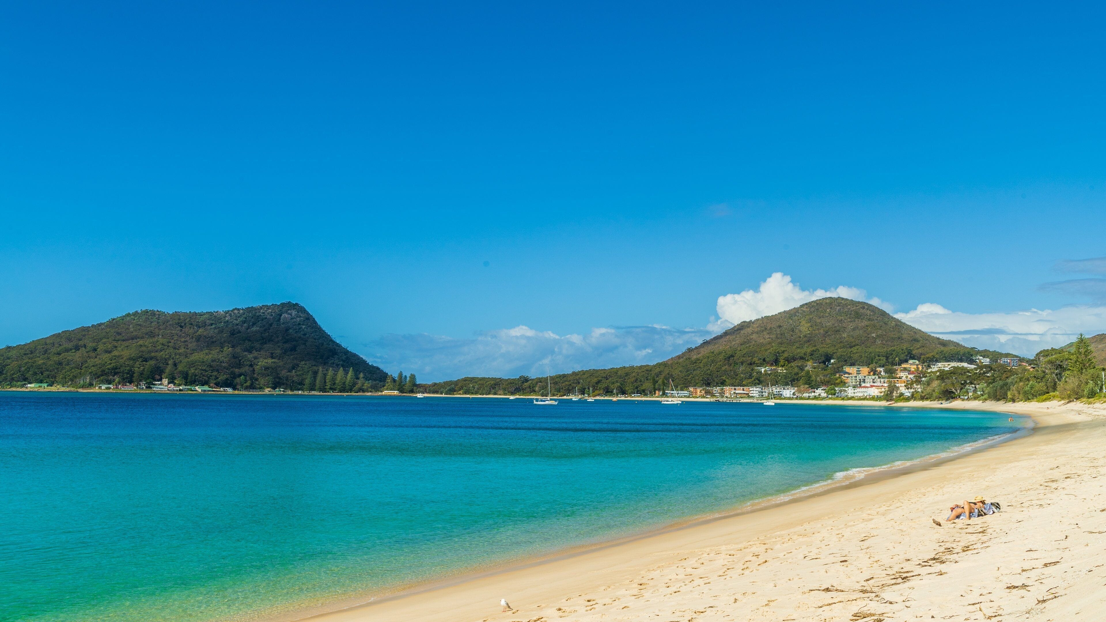 Een strand met helder turquoise water, een zandstrand en groene heuvels op de achtergrond.