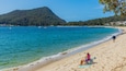 A beach with a mountain in the background, a person fishing, and a beach chair with a towel.