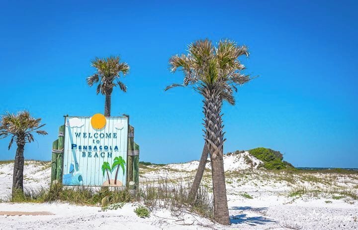 A sign welcoming visitors to Pensacola Beach with palm trees and a clear blue sky.