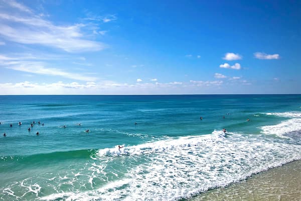 A beach with clear turquoise water, gentle waves, and a few people swimming and surfing.