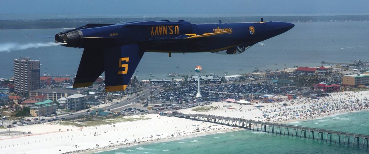 A blue and yellow airplane flying over a beach with a pier and a cityscape in the background.