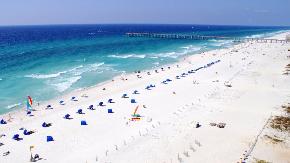 A beach with rows of blue umbrellas and a pier extending into the ocean.