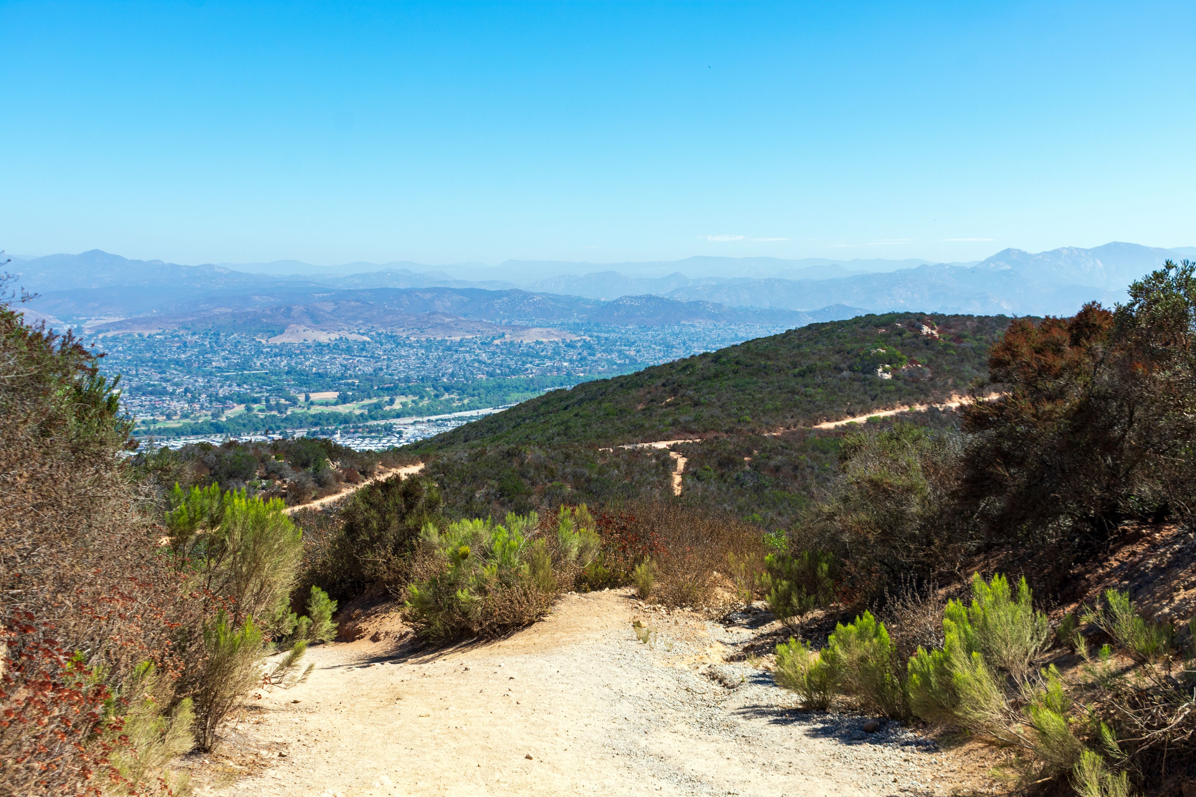 A winding dirt trail on Cowles Mountain in Mission Trails Regional Park offers expansive views of suburban sprawl and distant mountains under a clear blue sky