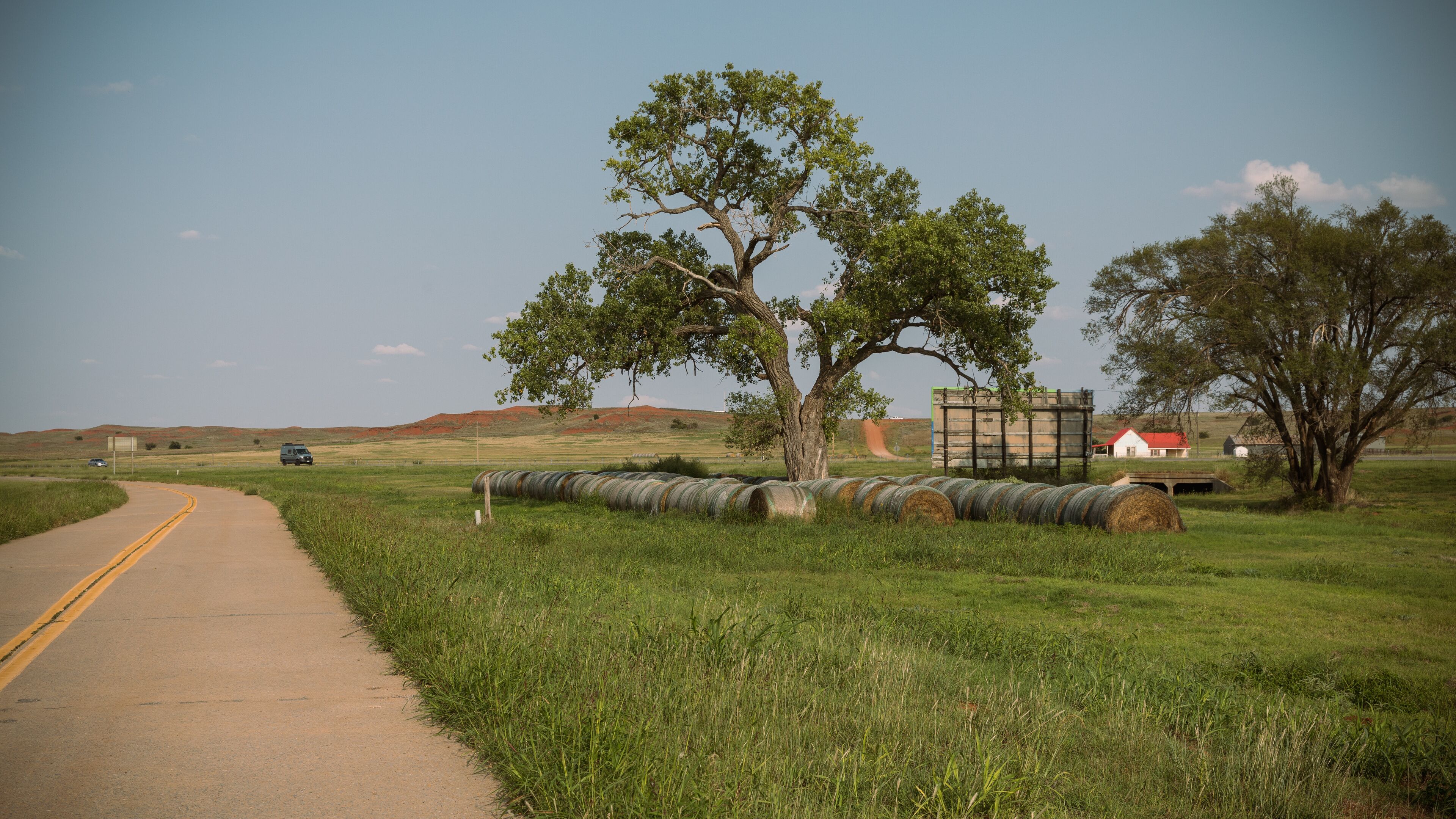 A rural road with a large tree, hay bales, and a red-roofed building in the distance.