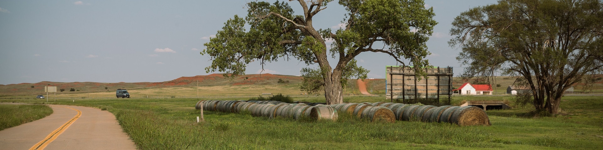 A rural road with a large tree, hay bales, and a red-roofed building in the distance.
