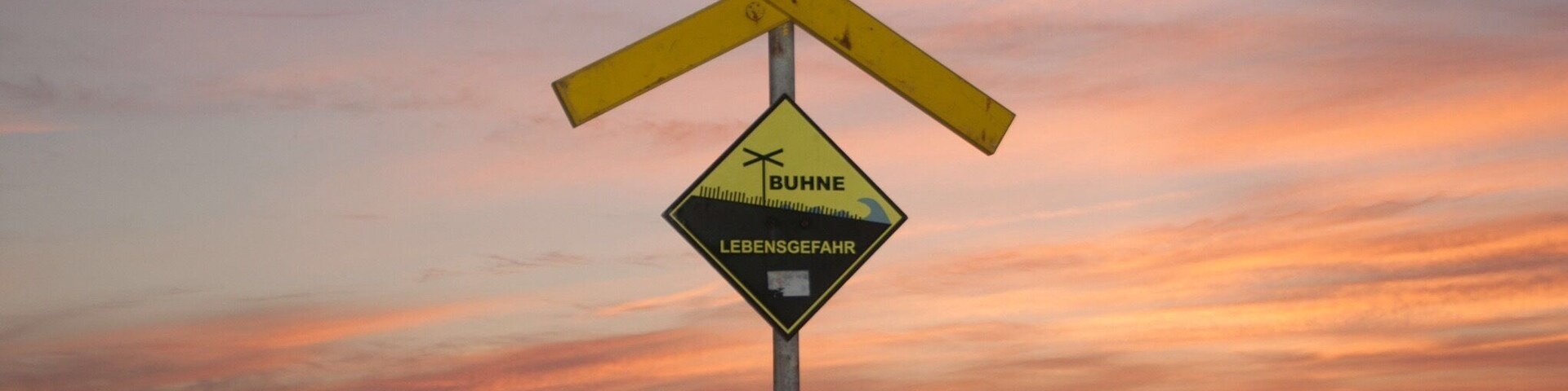 Señal de paso a nivel con la inscripción 'BUHNE' y 'LEBENSGEFAHR', sobre un cielo al atardecer.