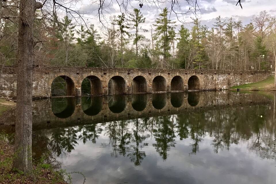 A stone bridge with multiple arches over a calm river, surrounded by trees and a cloudy sky.