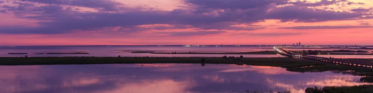 Un coucher de soleil sur une étendue d’eau, avec un pont et les lumières d’une ville lointaine.