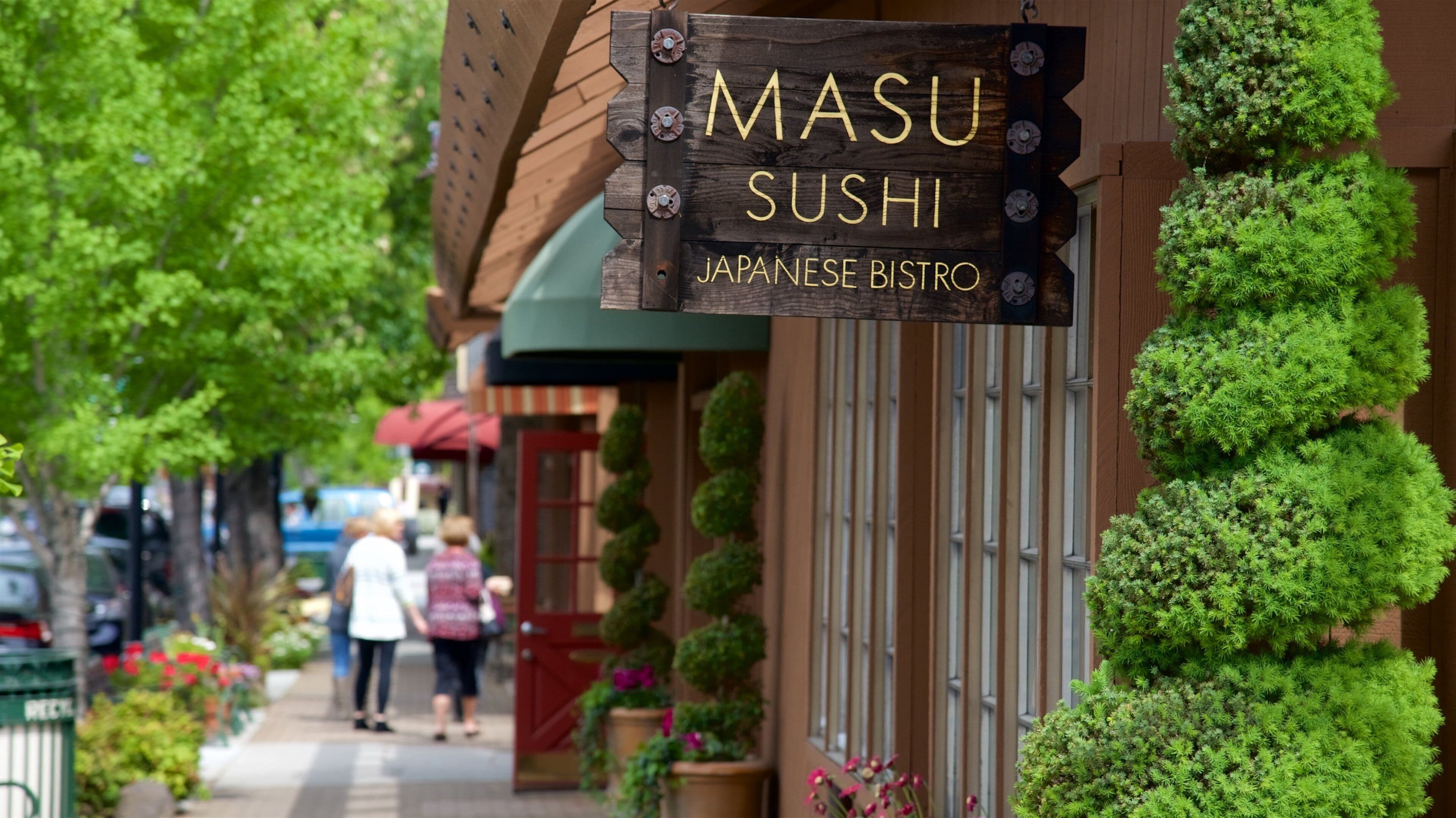 A wooden sign for Masu Sushi, a Japanese bistro, with green plants on the wall.