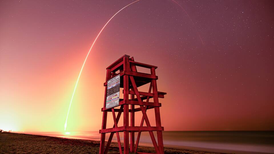 A red lifeguard tower on a beach at sunset with a visible rocket trail.