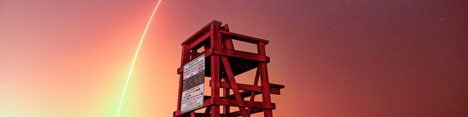 Una torre roja de salvavidas en una playa al atardecer, con el rastro visible de un cohete.