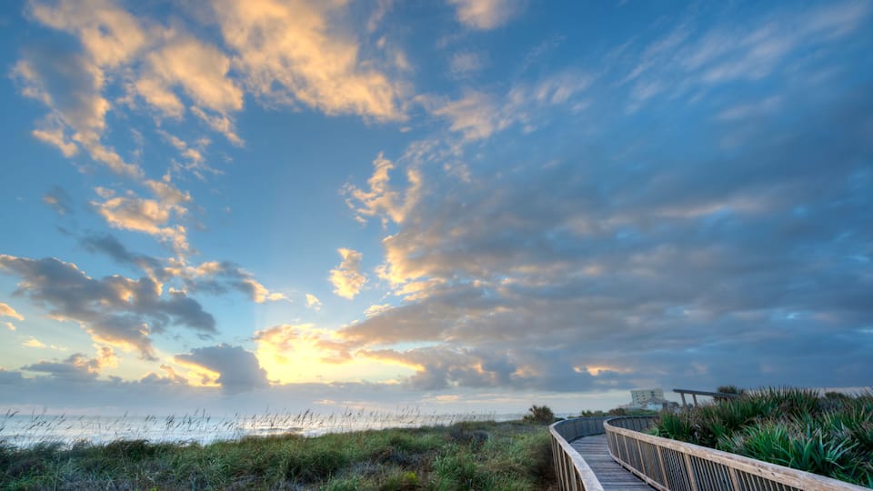 A wooden boardwalk along a coastal path with grassy dunes and a sunset sky.