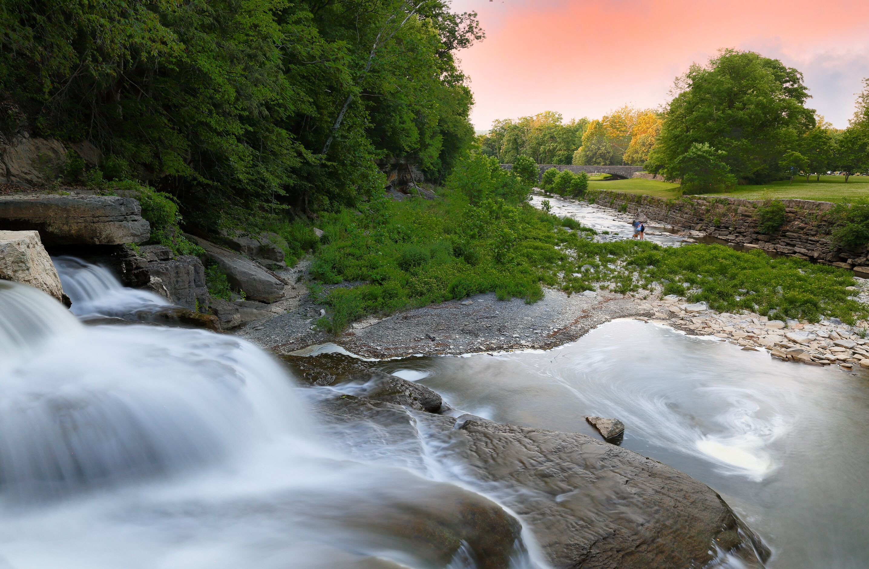 Un río serpentea por una zona boscosa con un puente de piedra al fondo.