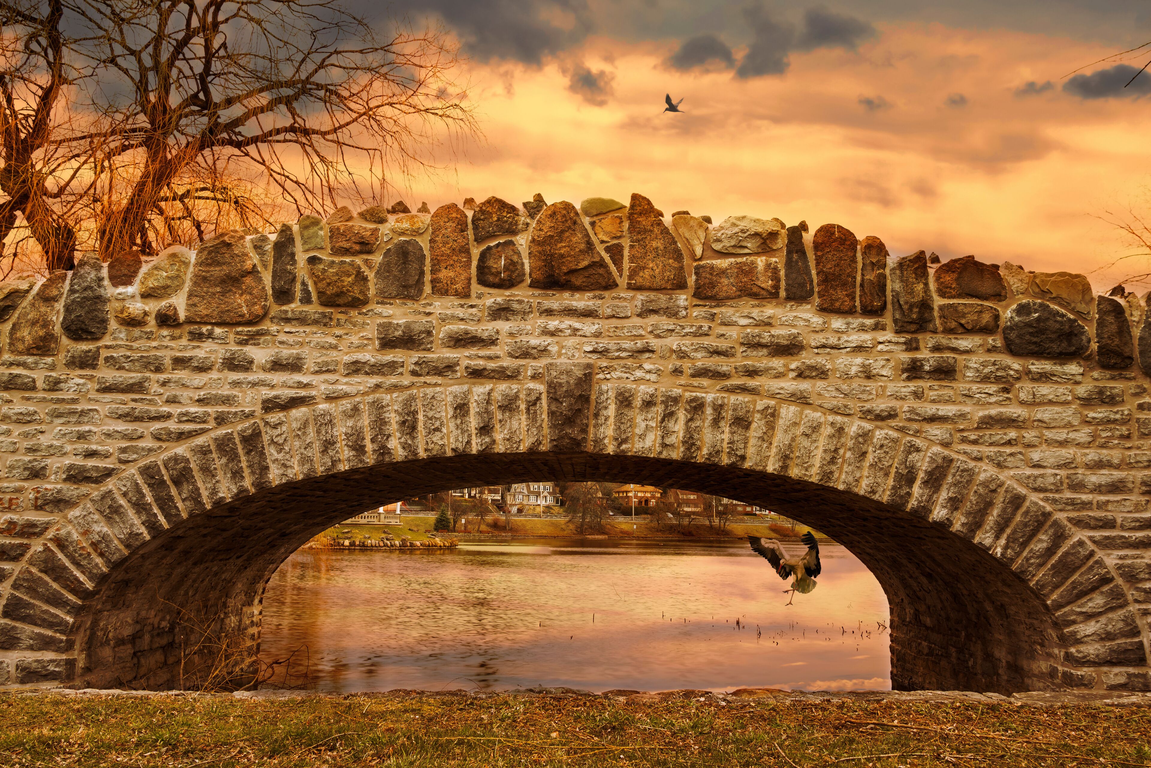 A stone bridge over a body of water with a reflection, surrounded by stone walls and a leafless tree.