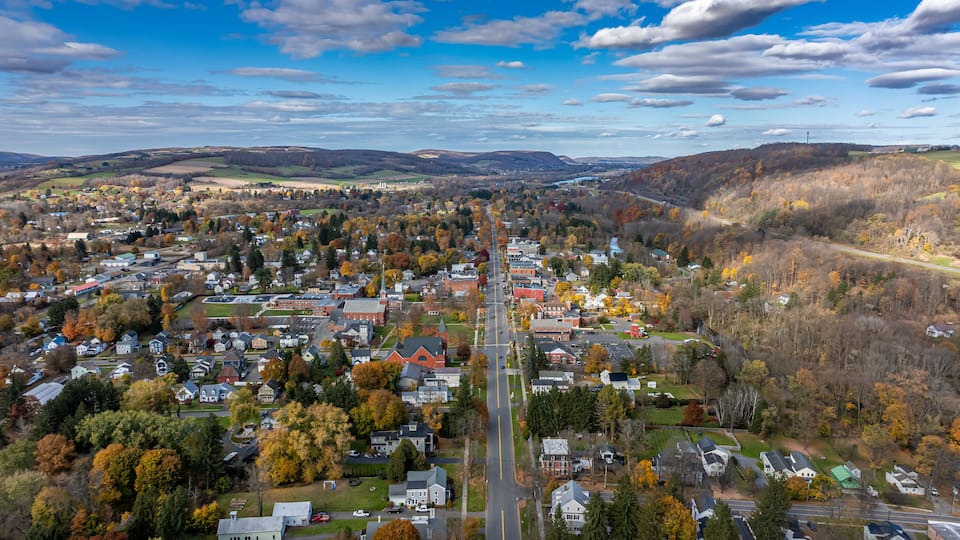 A town with a central road, surrounded by autumn-colored trees and rolling hills in the background.