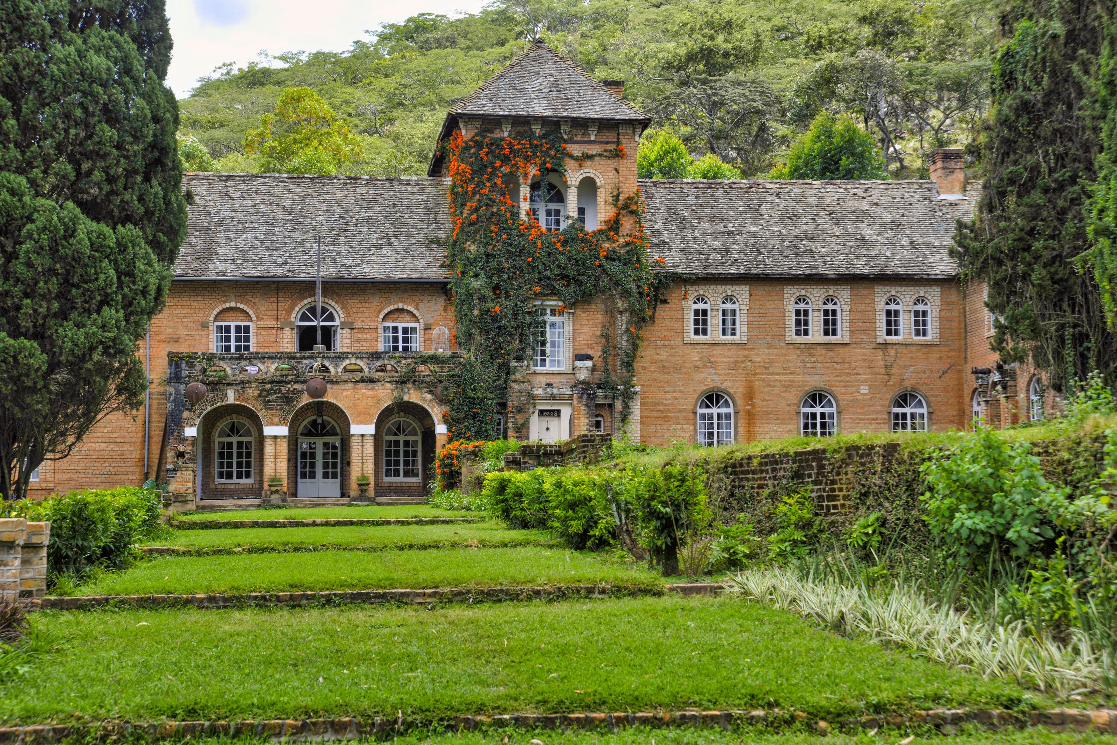 Un edificio histórico con ventanas de arco y una torre, rodeado de abundante vegetación y un jardín bien cuidado.