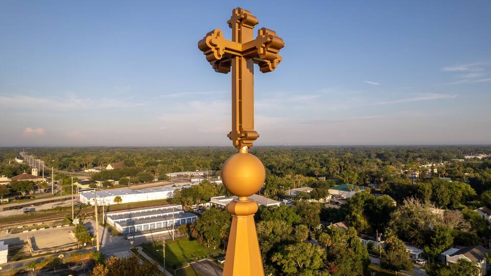 A golden cross atop a building with a view of a suburban area, including houses and a large white building.