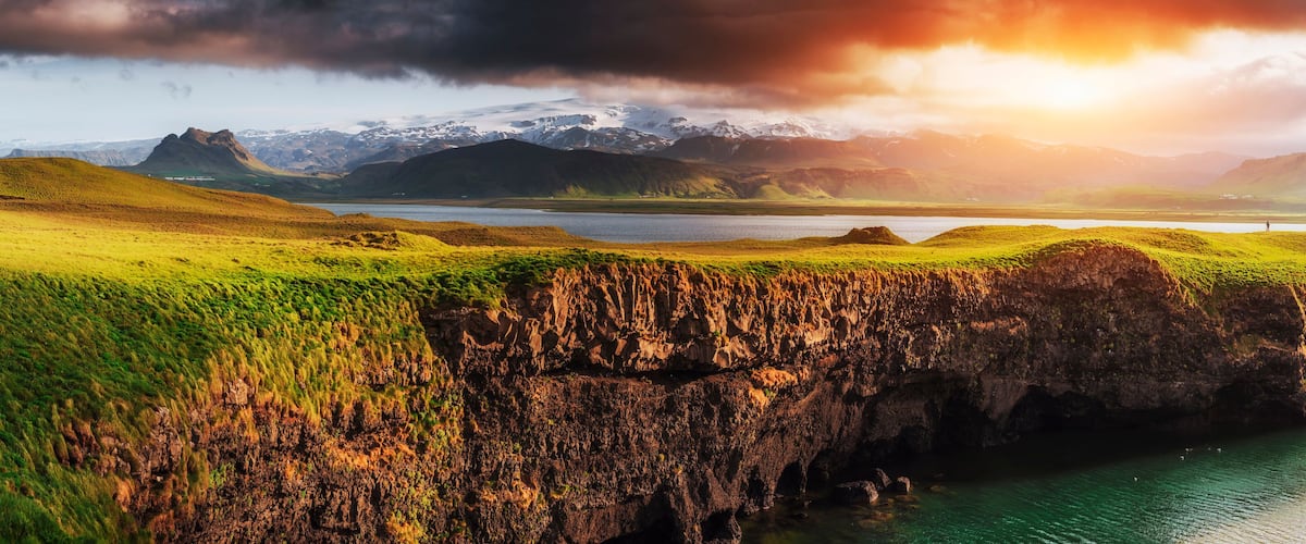 A coastal landscape with green hills, a rocky cliff, and a calm sea.