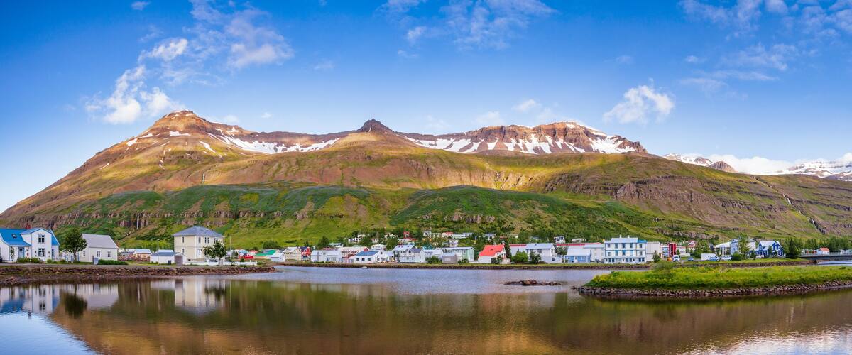 A coastal town with houses by a calm lake, surrounded by mountains with snow patches.