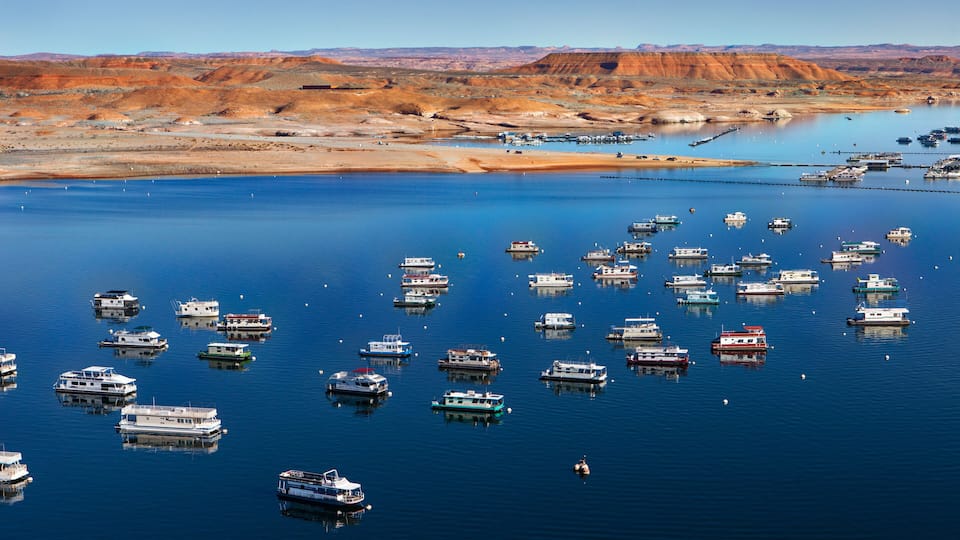A marina with numerous boats docked in calm waters, surrounded by a desert landscape with reddish-brown hills.