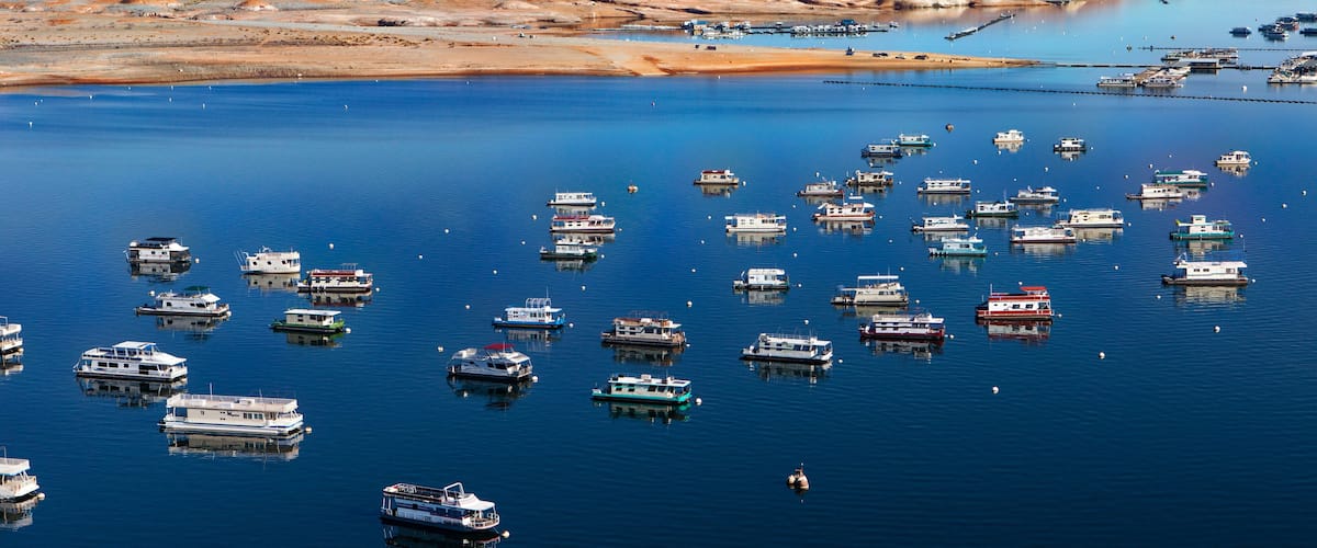 A marina with numerous boats docked in calm waters, surrounded by a desert landscape with reddish-brown hills.