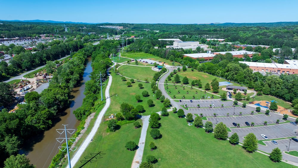 An aerial view of a park with a river, parking areas, and surrounding buildings.