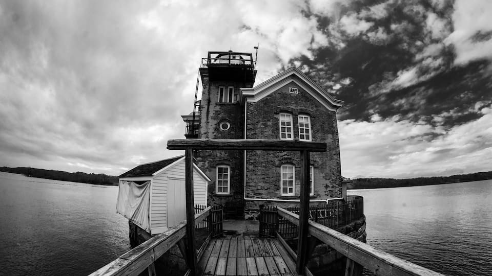 A wooden pier leading to a historic brick house with a tower, overlooking a calm lake.