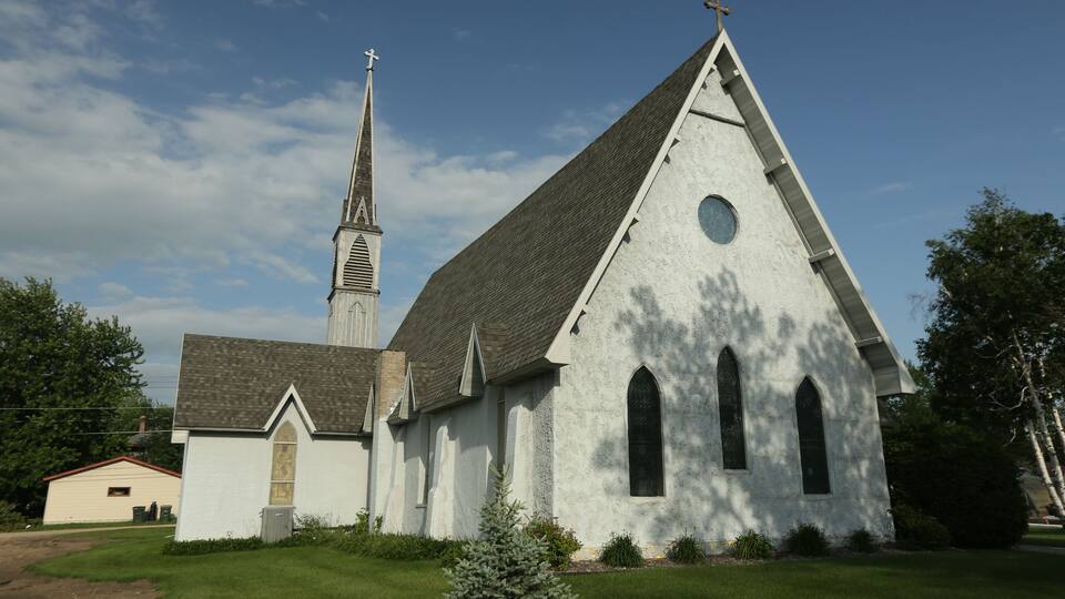 A white church with a steeple and a cross on top, surrounded by a well-maintained lawn and trees.