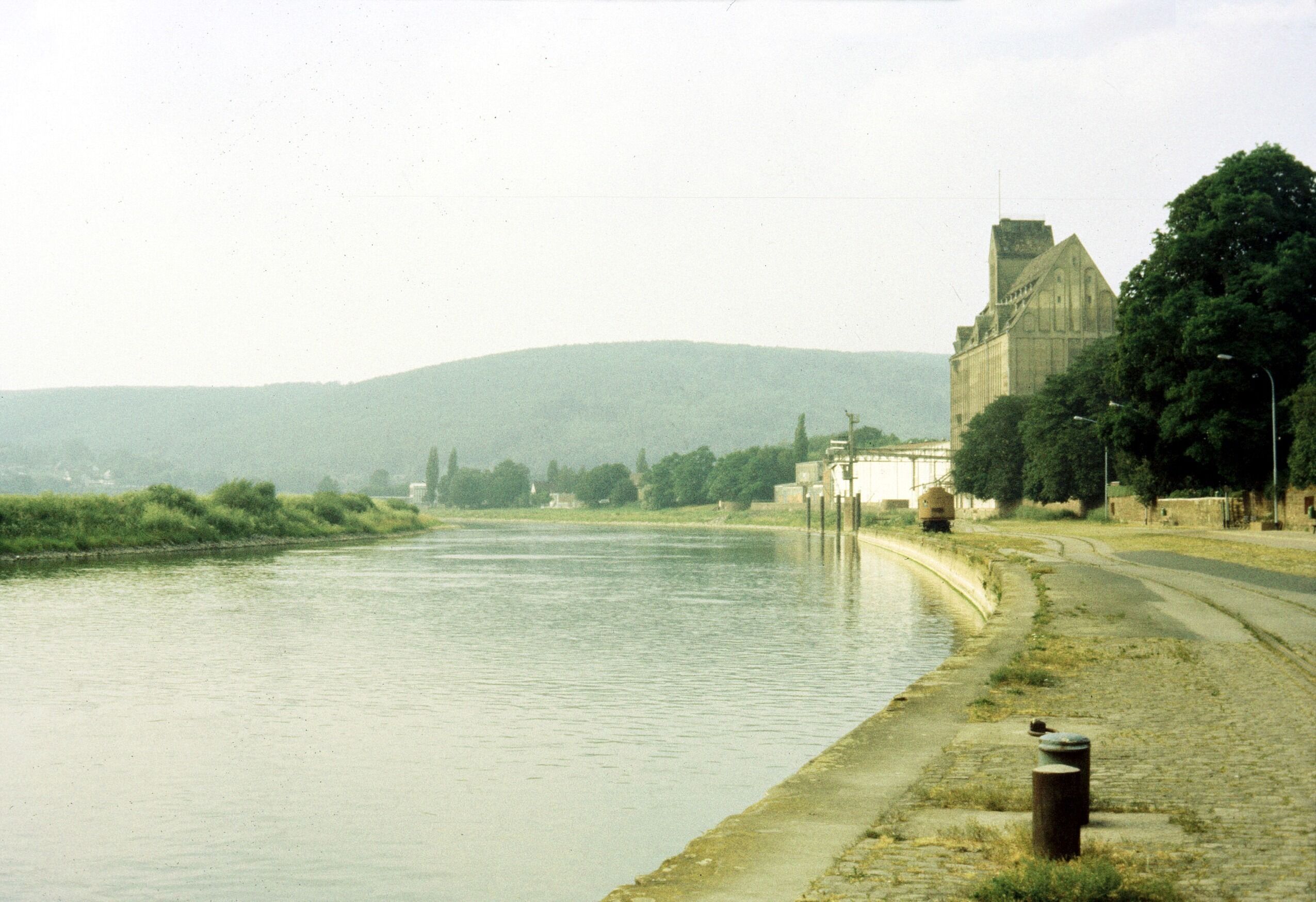A river with a stone embankment, a large building with a tower, and a hilly landscape in the background.