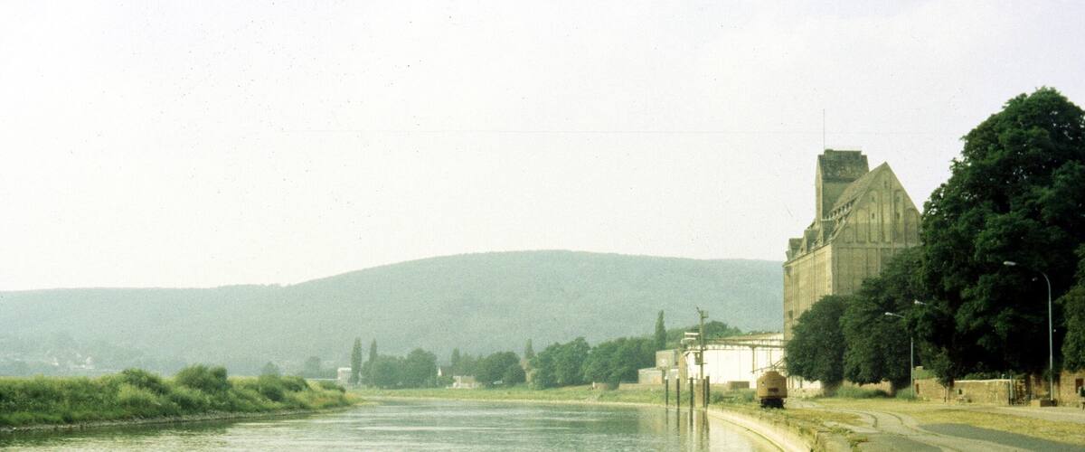 A river with a stone embankment, a large building with a tower, and a hilly landscape in the background.