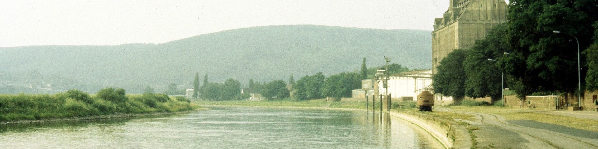 A river with a stone embankment, a large building with a tower, and a hilly landscape in the background.