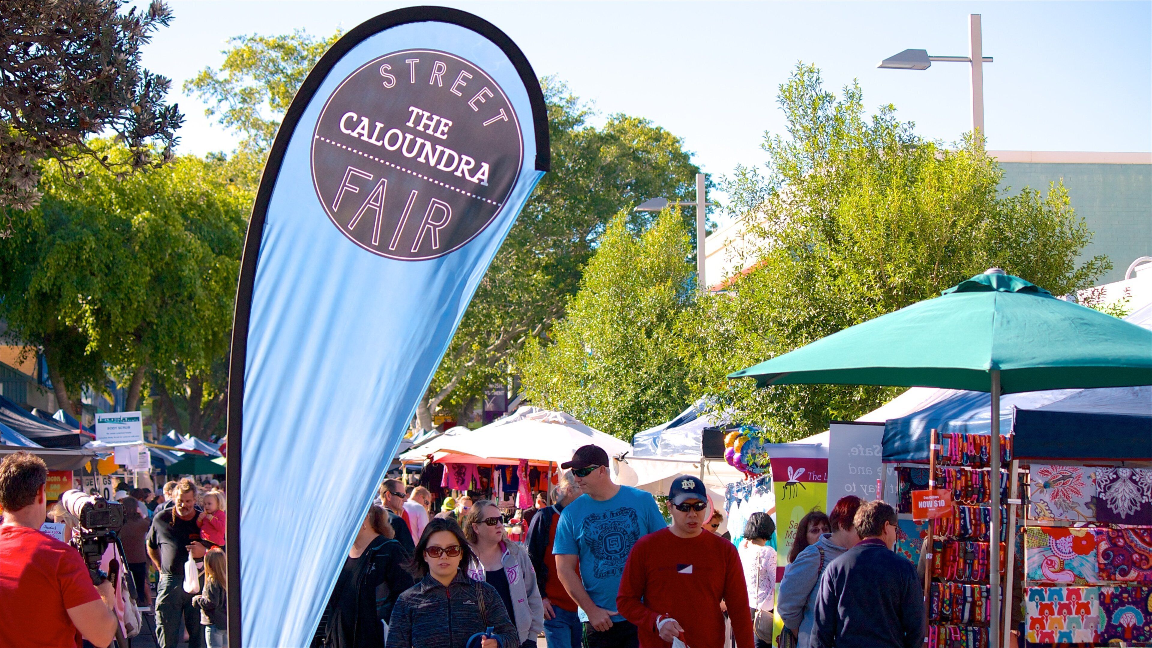 A street sign for The Caloundra Fair with a crowd of people and market stalls in the background.