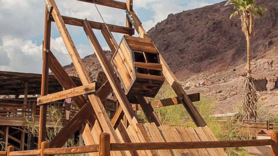 A wooden structure with a ladder and a wooden fence in a desert landscape with a palm tree.