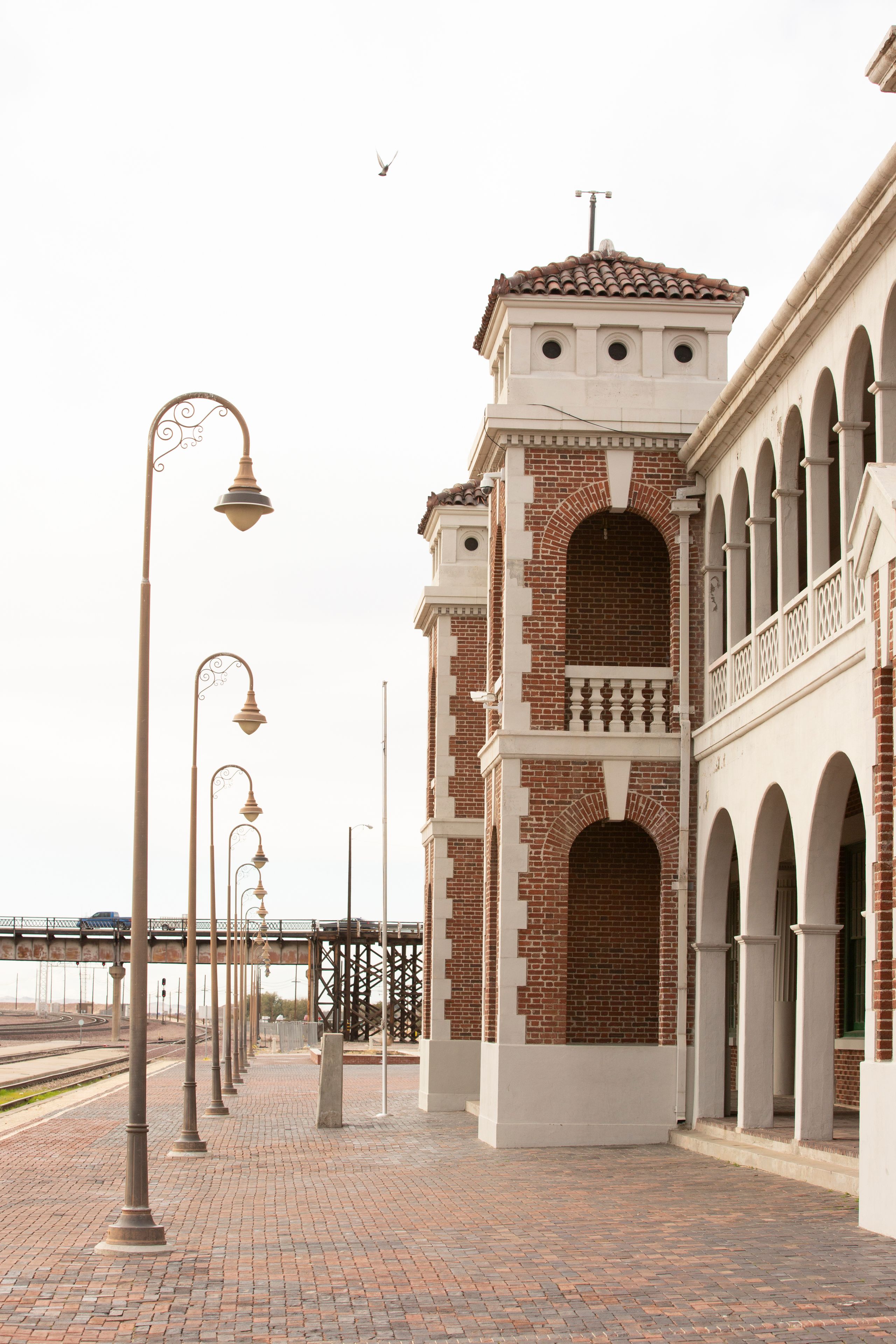 A brick building with arches and a tower, street lamps, and a bridge in the background.