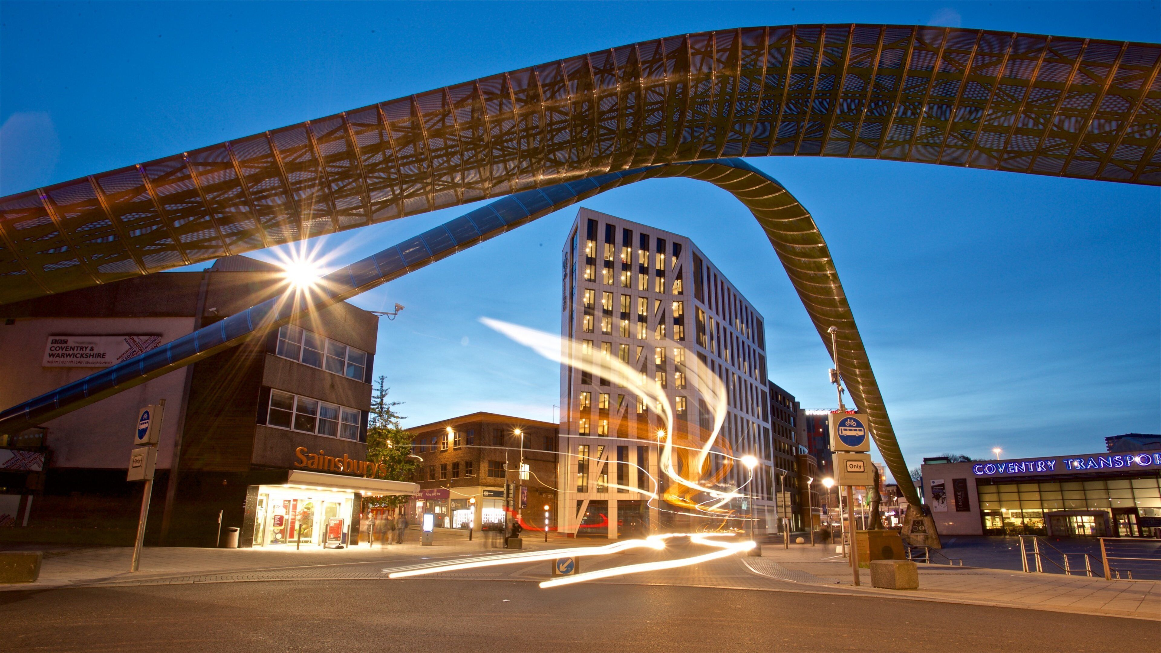 A city street at dusk with a prominent arch structure, a Sainsbury's store, and a Coventry & Warwickshire sign.
