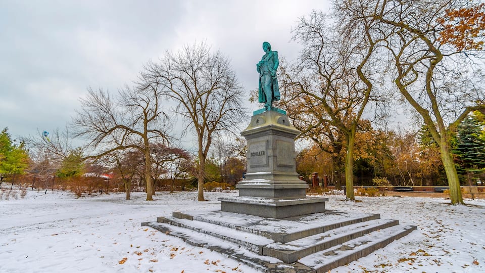 A statue of a man on a pedestal in a park with snow on the ground and bare trees.
