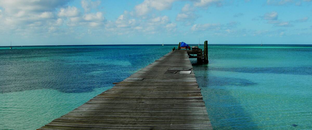 A wooden pier extending into a clear blue sea with boats in the distance.