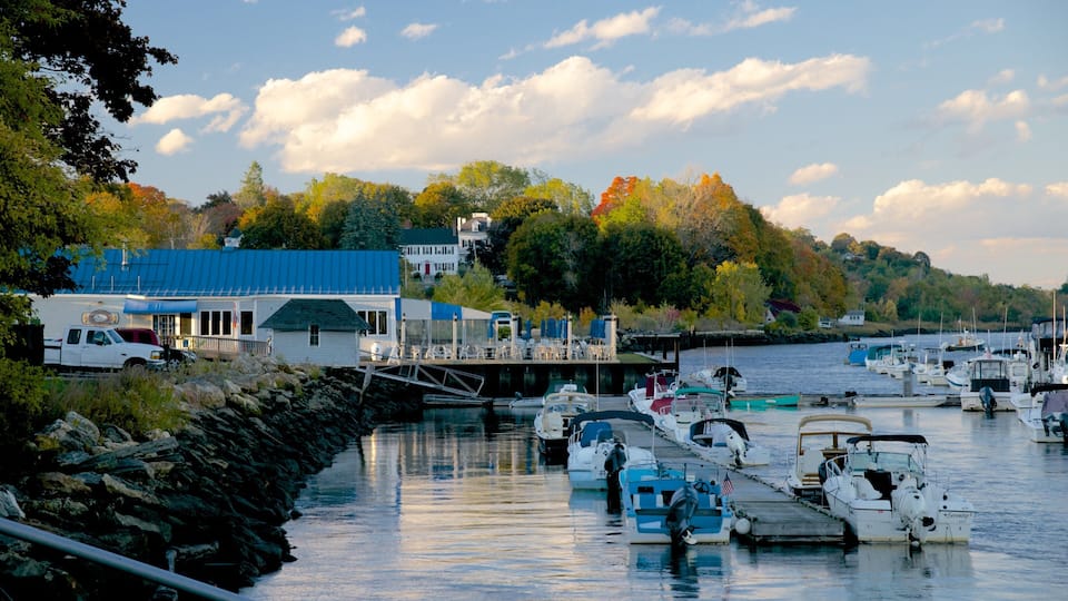 A marina with boats docked, a white building with a blue roof, and trees with autumn foliage in the background.