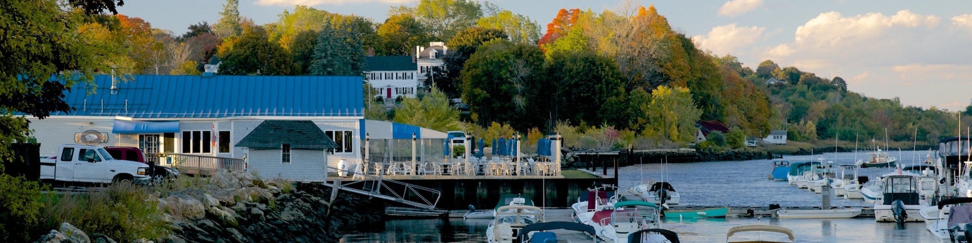 A marina with boats docked, a white building with a blue roof, and trees with autumn foliage in the background.