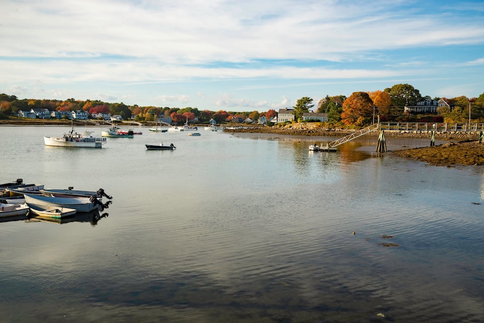 A calm lake with several boats docked at the pier, surrounded by houses and autumn-colored trees.