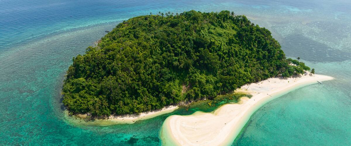 An aerial view of a tropical island with dense green forest, a sandy beach, and clear turquoise waters.