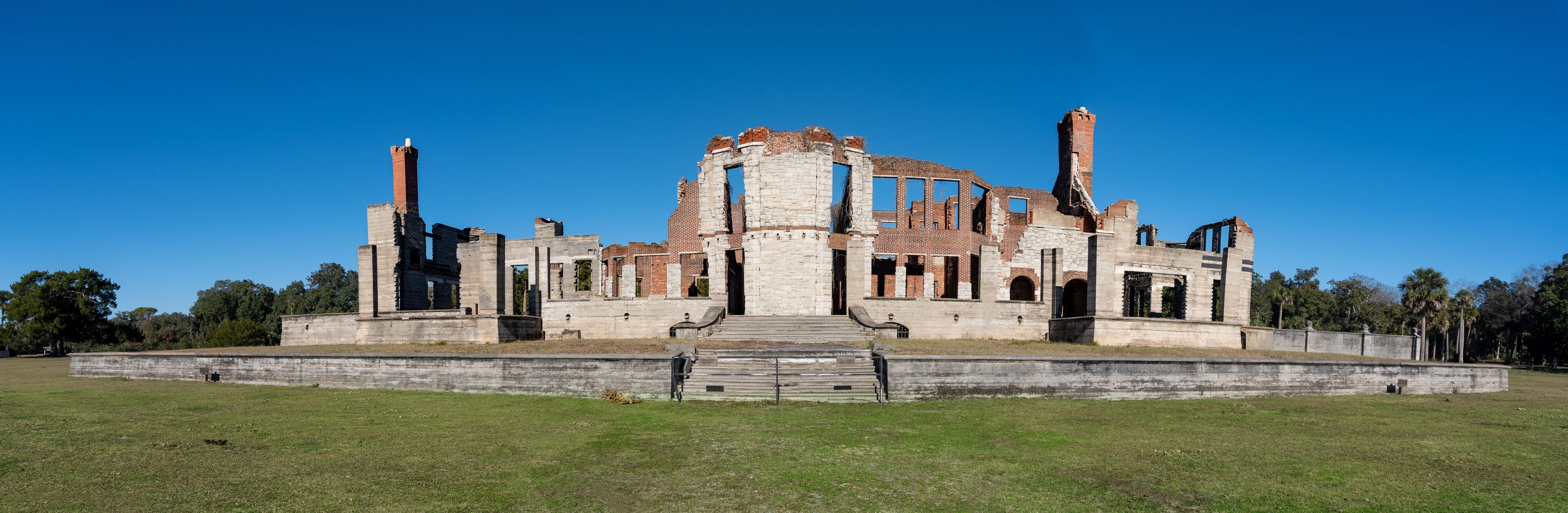 A large, historic stone building with multiple towers and arched doorways, surrounded by a grassy field and trees.