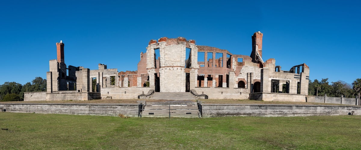 A large, historic stone building with multiple towers and arched doorways, surrounded by a grassy field and trees.