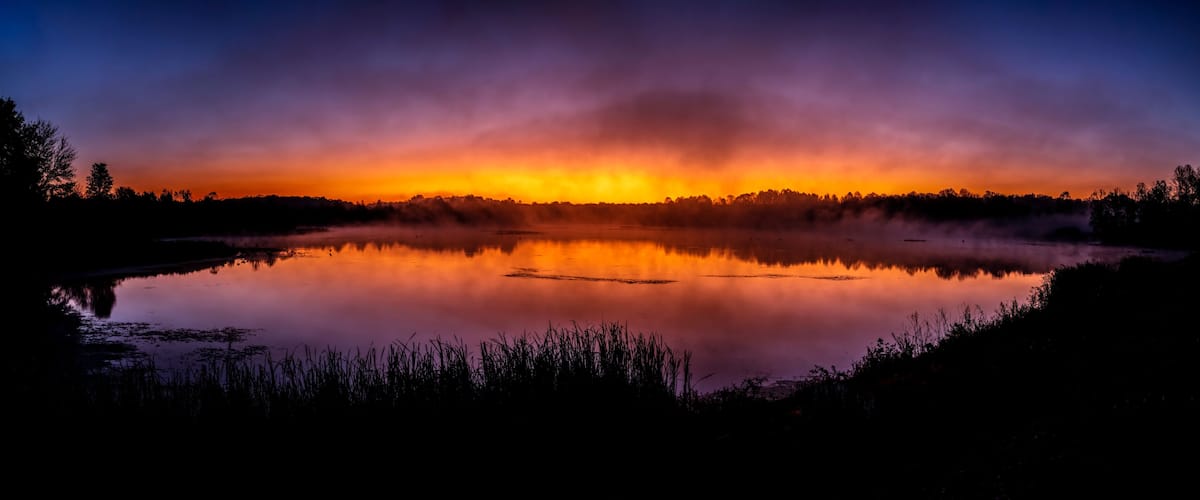 A serene lake at sunset with a reflection of the sky, surrounded by trees and vegetation.
