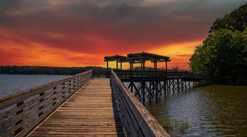 a long brown wooden bridge over the rippling blue waters of Lake Acworth surrounded by lush green trees and plants with powerful clouds at sunset at Cauble Park in Acworth Georgia USA