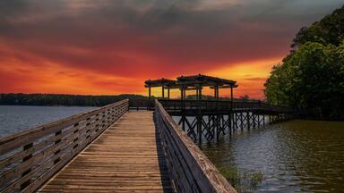 a long brown wooden bridge over the rippling blue waters of Lake Acworth surrounded by lush green trees and plants with powerful clouds at sunset at Cauble Park in Acworth Georgia USA