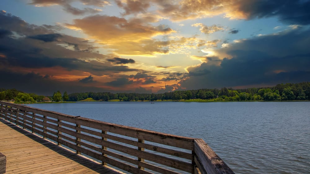 a brown wooden bridge over the silky green waters of Lake Acworth surrounded by lush green trees and plants with powerful clouds at sunset at Cauble Park in Acworth Georgia USA