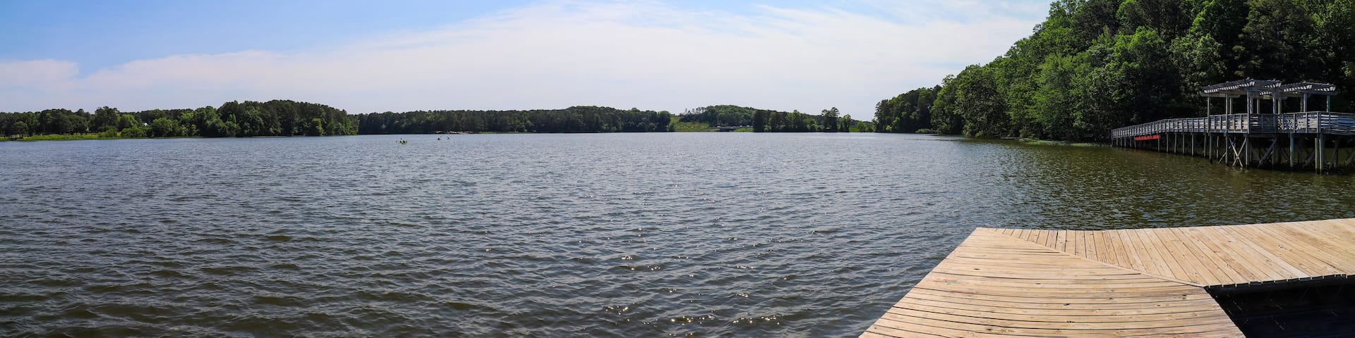 a panoramic shot of the rippling blue waters of Lake Acworth with a brown wooden bridge and dock surrounded by lush green trees, grass and plants with blue sky and clouds at Cauble Park in Acworth